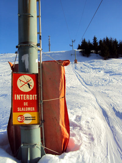 <Picture of the St Jean de Sixt ski area and interesting old sign>“>But before I get to that, look at this lovely sign! It’s one of a few dotted around the place and reveals the odd history of the resort. This jerky old drag lift is one of two lifts in the area. The other is a rope tow for beginners, while this one started life closer to the main road to La Clusaz back in 1962. The entire lift was moved to its current position in 1971, and I’m guessing the signs were not updated. This particular sign says that it’s forbidden to ski outside the tracks.</p>
<p>On the day that I went to the resort with a friend, there was only one other customer. He was using the beginner slope, but left soon after, and we had to wait for the man in charge of this longer lift to get back from his lunch break before he cranked it up for the afternoon rush. In fact, we were the afternoon rush. In two hours, nobody else arrived and the lift was due to close soon after!</p>
<p>The pistes from the top include a green, a blue and even a red. They’re all very short but lots of fun. My friend even tried a tree run through the dense forest with some success. Who knew there was off-piste right here in St Jean de Sixt? At the time of writing, there was even a whole web page devoted to the resort (no longer available), including a map and lots of photos.</p>
<p>So, why am I classing this place as an alternative to downhill winter sports? Because getting down isn’t the challenge at all: this drag lift —with a 62° slope half way up, a jumpy cable that sends you flying a few times during the ride up, and a flat section that means you have to leave the tracks despite the sign demanding you don’t — is the real sport. And so, I’m classing the ride on the drag lift as an alternative to downhill winter sports.</p>
<span id=